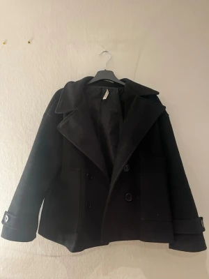 Svart kappa från Design by Si - Stilren svart kappa från Design by Si. Kappan har stora fickor och dekorativa knappar vid ärmsluten. Perfekt för dig som vill ha en klassisk och tidlös look. Fodrad insida för extra komfort. Har ett hål i armhålan i innertyget så inget som syns utanpå 