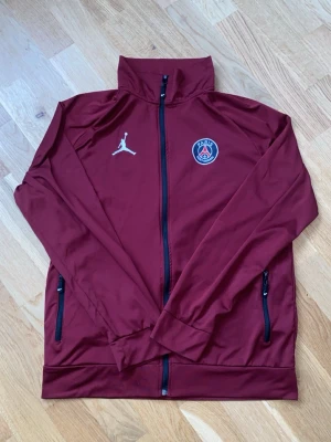 Vinröd PSG träningsjacka Jordan - Snygg vinröd träningsjacka från Jordan x PSG med dragkedja framtill och två fickor med svarta dragkedjor. Jackan har PSG-logga och Jumpman-logga på bröstet. Perfekt för träning eller som sportig streetwear.
