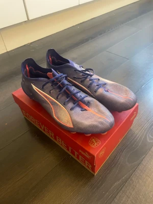 Puma lila fotbollsskor Elite/ultimate - Säljer ett par snygga fotbollsskor från Puma i lila med orange och vita detaljer. Detta är elites/ultimate alltså den dyraste och bösta  modell av denna färg, låg snörning och är tillverkade i ett lätt syntetmaterial för bästa känsla på planen. Perfekta för dig som vill sticka ut på matchen! Storleken är rätt stor i sig och skulle säga den passar 44.5 och 45 som bäst. Kostar 2700kr nypris