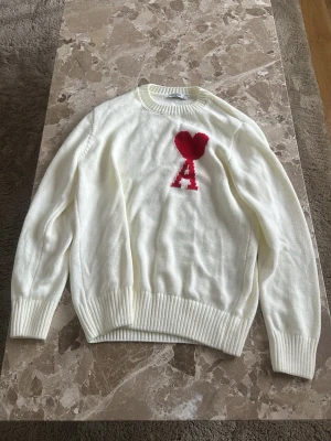 Vit stickad tröja från Ami - Säljer en vit stickad tröja från Ami med ett stort rött hjärta och bokstaven A på bröstet. Tröjan har rund halsringning och ribbstickade muddar vid ärmslut och nederkant. Perfekt för dig som gillar stilrena detaljer och coola loggor.