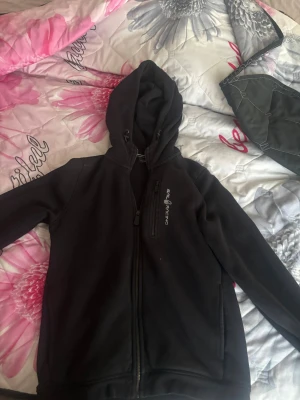 Svart hoodie från Sea Racing - Svart hoodie med dragkedja från Sea Racing. Tröjan har huva med dragsko, två sidofickor och en bröstficka med dragkedja. Perfekt för en avslappnad och sportig stil. Materialet ser ut att vara mjukt och bekvämt, troligen bomull eller fleece.