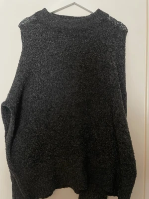 Mörkgrå stickad tröja - Mysig mörkgrå stickad tröja med lös passform och ribbade muddar. Tröjan har en enkel design utan synliga detaljer eller mönster och är tillverkad i ett mjukt, fluffigt material som känns skönt mot huden. Perfekt för kyliga dagar.