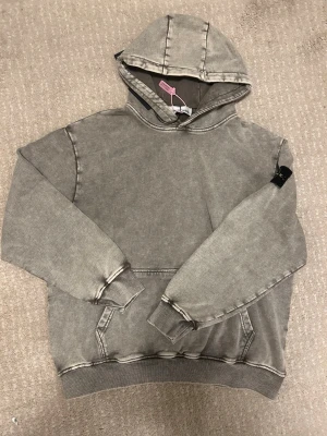 Grå hoodie från Stone Island - Säljer en grå hoodie från Stone Island med klassisk patch på vänster ärm. Hoodien har en tvättad vintage-look, stor huva och känguruficka framtill. Tillverkad i mjuk bomull och har ribbade muddar vid ärmslut och nederkant. Perfekt för en avslappnad streetwear-stil.