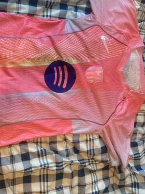Rosa FC Barcelona matchtröja Nike - Snygg rosa FC Barcelona matchtröja från Nike med klubbmärke och Spotify-logga på bröstet. Tröjan har korta ärmar, rund hals och är tillverkad i ett lätt, ventilerande funktionsmaterial. Perfekt för fotboll eller träning.