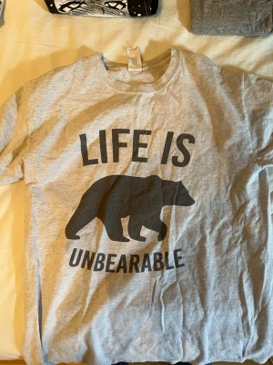 Grå t-shirt med björntryck - Säljer en grå t-shirt med svart tryck av en björn och texten 'LIFE IS UNBEARABLE'. T-shirten har rund hals och korta ärmar. Perfekt för dig som gillar humoristiska motiv och enkel stil.
