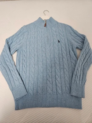 Ljusblå kabelstickad tröja Polo Ralph Lauren - Snygg ljusblå kabelstickad tröja från Polo Ralph Lauren med halv dragkedja och hög krage. Tröjan har ribbade muddar och nederkant samt den klassiska broderade loggan på bröstet. Perfekt för lager på lager och en chill look.