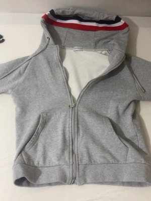 Grå Moncler hoodie med dragkedja - Snygg grå hoodie från Moncler med dragkedja framtill och två fickor. Huvan har en röd, vit och marinblå randig kant som ger en sportig vibe. Tillverkad i mjukt bomullsmaterial, perfekt för chill dagar. Klassisk passform och stilren design. Har används några gånger bara.