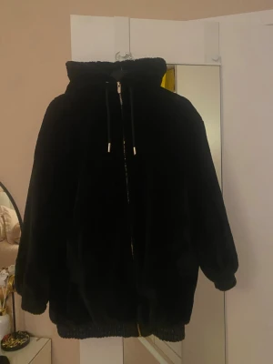 Svart teddyjacka med huva - Mysig svart teddyjacka med huva och dragkedja framtill. Jackan har oversized passform, ribbade muddar vid ärmslut och nederkant samt snörning i huvan. Perfekt för kalla dagar när du vill vara både varm och stilren.