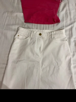 Vit jeanskjol med fickor - Säljer en vit jeanskjol med klassisk femficksdesign och knapp i midjan. Kjolen har en rak passform och är tillverkad i ett stadigt bomullstyg. Perfekt att matcha med en färgglad topp för en fräsch look. Toppen ingår till kjolen🌸