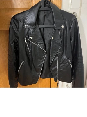 Svart biker skinnjacka med dragkedjor - Cool svart skinnjacka i bikerstil med silvriga dragkedjor och detaljer. Jackan har bred krage, snedställd dragkedja framtill och quiltade partier på ärmarna. Perfekt för en edgy look och passar till många outfits. 