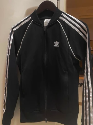 Svart Adidas zip-tröja XS - Cool svart zip-tröja från Adidas med klassiska vita ränder längs ärmarna och Adidas logga på bröstet. Tröjan är tillverkad av Primeblue-material och har ribbade muddar samt fickor på sidorna. Perfekt för en sportig look.