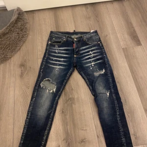 Dsquared2 mörkblå  jeans - Snygga mörkblå jeans från Dsquared2 med slitningar och hål på benen för en cool, edgy look. Jeansen har klassisk femficksdesign, kontrastsömmar och Dsquared2-logga på bakfickan. Passformen är smal och materialet är robust denim.