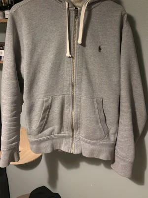 Grå hoodie från Polo Ralph Lauren - Grå zip-hoodie med vit dragsko och Polo Ralph Laurens klassiska broderade logga på bröstet. Mjuk bomullskänsla, två fickor framtill och ribbade muddar. Perfekt för en chill och stilren look.