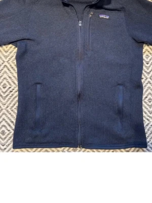 Mörkblå fleecejacka från Patagonia - Snygg mörkblå fleecejacka från Patagonia med hel dragkedja, bröstficka och två sidofickor. Jackan har hög krage och diskret logga på bröstet. Perfekt för lager-på-lager och chill vardagsstil.
