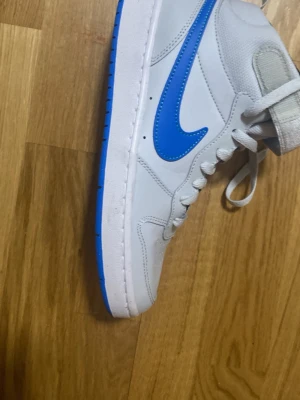 Nike vita och blå sneakers - Nike sneakers i vitt med blå detaljer och sula. Skon har en klassisk hög siluett med blå swoosh på sidan och texten 'ALL DAY PLAY' under sulan. Ovandelen är i syntetmaterial och mesh för extra komfort. Perfekt för dig som gillar sportig och clean stil.