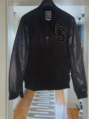 Svart bomberjacka från G-Star RAW - Snygg svart bomberjacka från G-Star RAW med broderad GS-logga på bröstet och stor Gstar-text på ryggen. Jackan har glansigt material, ribbade muddar och dragkedja framtill. Perfekt för dig som vill ha en streetwear-look.