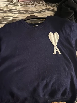 Marinblå stickad tröja från Ami Paris - Säljer en svart stickad tröja från Ami Paris med deras ikoniska vita hjärta och bokstaven A broderat på bröstet. Tröjan har rund halsringning och långa ärmar. Perfekt för dig som gillar stilrena och trendiga plagg.
