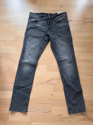 Grå jeans från Jack & Jones - Säljer ett par grå jeans från Jack & Jones med klassisk femficksdesign och knappgylf. Jeansen har raka ben och är tillverkade i denim med lätt tvättad look. Perfekta för dig som gillar en stilren och avslappnad vibe.
