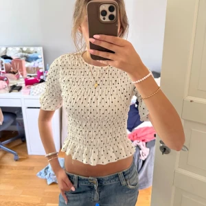 Prickig blus från Gina Tricot - Supersöt vit blus med svarta prickar från Gina Tricot. Modellen är croppad med smock över hela toppen, volangkant nertill och puffiga korta ärmar. 