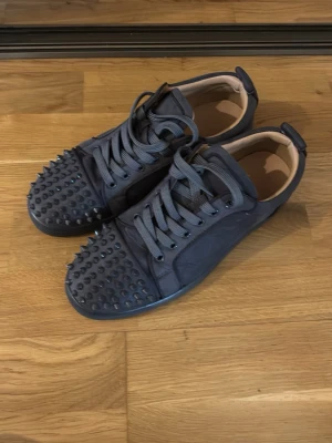 Christian Louboutin gråa med spikar - Säljer ett par gråa sneakers från Christian Louboutin med matta nitar på tån och sidorna. Skorna har snörning, låg profil och är tillverkade i mocka med läderdetaljer. Två spikar är lite lösa men går enkelt att limma fast. Knappt använda! Storlek 42