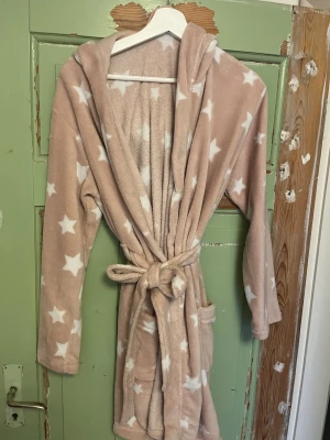 Beige morgonrock med stjärnor och huva - Mysig beige morgonrock med vita stjärnor, tillverkad i mjuk fleece. Modellen har huva, knytskärp i midjan och två praktiska fickor. Storlek XS. Hör av er om ni vill ha fler bilder. Priset kan alltid diskuteras!