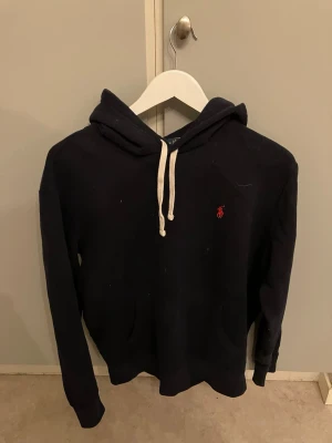 Mörkblå hoodie från Ralph Lauren - Snygg mörkblå hoodie från Ralph Lauren med vit dragsko och den klassiska röda loggan broderad på bröstet. Hoodien har en stor huva och känguruficka framtill. Perfekt för en avslappnad och stilren look.