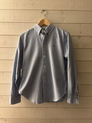 Blå skjorta från Ralph Lauren - Klassisk ljusblå skjorta från Ralph Lauren med button-down krage och broderad logga på bröstet. Skjortan har långa ärmar och knappar framtill. Perfekt till en clean och stilren look.