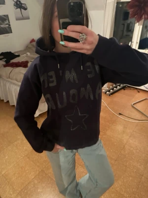 Mörkblå hoodie med stjärna och text - Säljer en mörkblå hoodie med huva och snören. På framsidan finns en stor stjärna och texten 'JE M'EM AMOUR' i glittrigt material. Från Ginatricot!!!