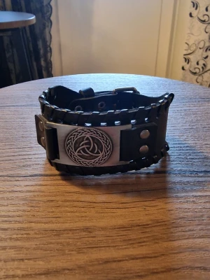 Armband vintage triangel - Svart läderarmband med ett detaljerat triangel i metall. Armbandet har justerbara tryckknappar för att passa olika handledsstorlekar. Storlek 27,5 cm långt. 