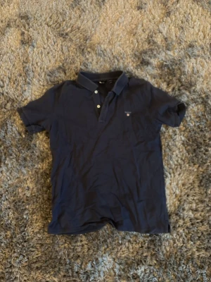 Mörkblå pikéskjorta från Gant - Säljer en mörkblå pikéskjorta från Gant med klassisk krage och knappar framtill. Skjortan är tillverkad i mjuk bomull och har korta ärmar, perfekt för en clean och stilren look.