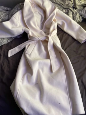 Ljusbeige kappa med skärp - Stilren ljusbeige kappa med bred krage och tillhörande midjeskärp. Kappan har långa ärmar, två sidofickor och stängs med knappar framtill. Materialet ser ut att vara mjukt och något tjockare, perfekt för kyligare dagar. Kappan är väldigt lång jag är 160 och den är till mina vader. Kappan är helt oanvänd då jag köpte fel. PRIS KAN DISKUTERAS 