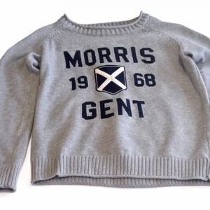 Grå stickad tröja Morris Gent - Snygg grå stickad tröja från Morris Gent med mörkblå text och ett broderat emblem framtill. Tröjan har rund hals, ribbstickade muddar och nederkant. Perfekt för en avslappnad men stilren look. Tröjan är vintage och säljs ej mer. Skicket är 9/10 och sällan använd. Priset är ej hugget i sten kan sänka vid snabb affär!