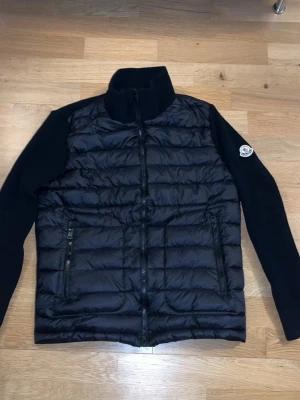 Svart pufferjacka från Moncler - Snygg svart pufferjacka från Moncler med quiltad front och stickade ärmar. Jackan har hög krage, två dragkedjeförsedda fickor och Moncler-logga på ena ärmen. Perfekt för kalla dagar och ger en stilren look.