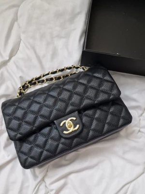 Svart Chanel quiltad handväska - Klassisk svart quiltad handväska från Chanel med guldfärgade detaljer och kedjerem. Väskan har det ikoniska CC-spännet framtill och är tillverkad i läder med struktur. Perfekt för dig som vill ha en tidlös och lyxig accessoar. Köpte utomlands. Tappade bort påsen men lådan följa med. 