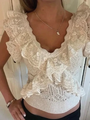 Beige spetsblus med volang från Zara - Supersöt beige blus från Zara med djup v-ringning och stora volangdetaljer framtill. Blusen är ärmlös och har ett romantiskt spetsmönster som ger en bohemisk vibe. Perfekt att styla med jeans eller kjol för en trendig look.