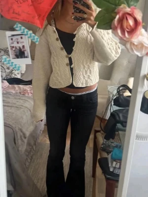 Vit kabelstickad kofta med knappar - Säljer en vit kabelstickad kofta med stora svarta knappar framtill. Koftan är croppad, har lång ärm och ett snyggt mönster med flätstickning både fram och bak. Perfekt att slänga över ett linne för en chill look.