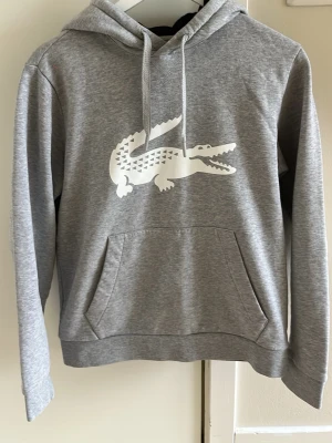 Grå hoodie från Lacoste med logga - Snygg grå hoodie från Lacoste med stor vit krokodil-logga på bröstet. Hoodien har huva med dragsko och en stor magficka framtill. Tillverkad i mjukt sweatshirtmaterial som är skönt mot huden. Perfekt för en avslappnad och sportig stil.