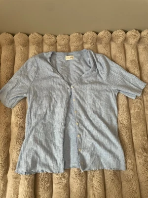 Blå ribbad blus - Säljer en ljusblå ribbad blus från Weekday med v-ringning och knappar framtill. Blusen har korta ärmar och är tillverkad i ett mjukt material som känns skönt mot huden. Perfekt att styla över ett linne eller t-shirt.