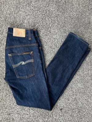 Mörkblå jeans från Nudie Jeans - Snygga mörkblå jeans från Nudie Jeans med klassisk femficksdesign och kontrastsömmar. Jeansen har smal passform och ikonisk broderad bakficka. Perfekta för dig som gillar stilrena och tidlösa jeans i denim.
