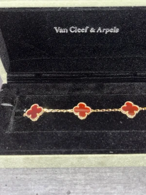 Van Cleef & Arpels Alhambra armband - Exklusivt armband från Van Cleef & Arpels med fyra fyrklöverformade hängen i röd sten, inramade av guldfärgade detaljer. Kedjan är tunn och elegant i guldton. Perfekt för dig som gillar lyxiga och ikoniska smycken med unik design.