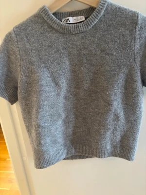 Grå kortärmad stickad tröja Zara - Mysig grå stickad tröja från Zara med korta ärmar och ribbade detaljer vid hals, ärmslut och nederkant. Tröjan har en enkel, rak passform och är tillverkad i ett mjukt material som känns skönt mot huden. Perfekt för lager på lager och en clean look.