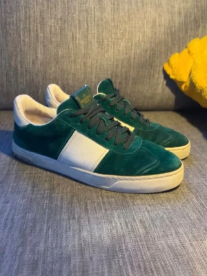 Gröna sneakers från valentino  - Snygga gröna sneakers från valentino i mocka med vita detaljer och vit sula. Skorna har en klassisk låg siluett och svarta skosnören. Perfekta för dig som vill ha en clean och stilren look med en twist av färg.