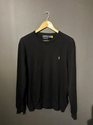 Svart tröja från Polo Ralph Lauren - Klassisk svart tröja från Polo Ralph Lauren med rund halsringning och diskret broderad logga på bröstet. Tillverkad i mjukt material och har ribbade muddar vid ärmslut och nederkant. Perfekt för en stilren look.