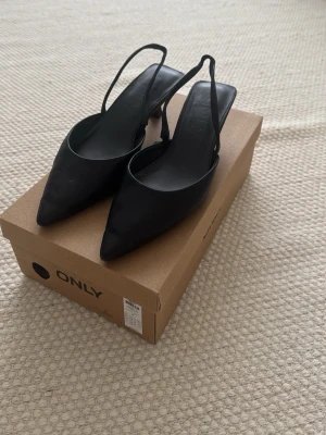 Svarta slingback pumps från ONLY - Stilrena svarta pumps från ONLY med spetsig tå och smal slingback-rem. Skorna har låg klack och är tillverkade i ett slätt material som ger en elegant look. Perfekta för dig som vill ha en klassisk och trendig sko till garderoben.