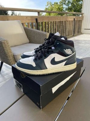 Air Jordan 1 Mid svart/vit - Snygga Air Jordan 1 Mid sneakers från Nike i svart och vitt läder med klassisk logga på sidan. Skorna har svart swoosh, svarta skosnören och vit mellansula. Perfekta för dig som gillar streetwear och vill ha en ikonisk look.