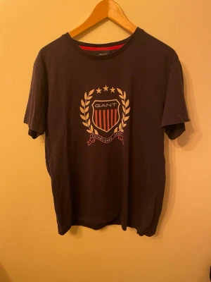 Svart GANT t-shirt med tryck - Snygg svart t-shirt från GANT med stort tryck framtill i form av ett emblem med guldiga detaljer och röda ränder. Klassisk rund halsringning och korta ärmar. Tillverkad i mjuk bomull som känns skön mot huden. Perfekt för en stilren och sportig look.