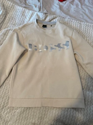 Beige sweatshirt från BOSS - Snygg beige sweatshirt från BOSS med silverfärgad logotyp på bröstet. Tröjan har rund halsringning, långa ärmar och ribbade muddar vid ärmslut och nederkant. Perfekt för en avslappnad och trendig look.