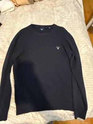 Mörkblå stickad tröja från Gant - Klassisk mörkblå stickad tröja från Gant med rund halsringning och diskret logga broderad på bröstet. Tröjan har långa ärmar och ribbade muddar vid ärmslut och nederkant. Perfekt basplagg för garderoben.