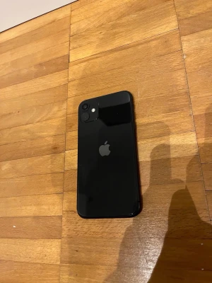 Svart iPhone 11 64GB - Säljer en svart iPhone 11 från Apple med stilren glasbaksida och rundade kanter. Mobilen har dubbla kameror på baksidan och den klassiska Apple-loggan. Perfekt för dig som vill ha en snygg och modern smartphone.