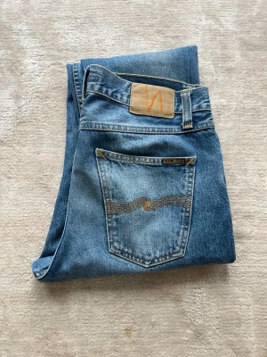 Archive Nudie Jeans - Säljer ett par klassiska blå jeans från Nudie Jeans med rak passform och fem fickor. Jeansen har en snygg tvättad look, kontrastsömmar och Nudie-loggan på bakfickan. Tillverkade i slitstark denim, perfekta för en avslappnad stil.
